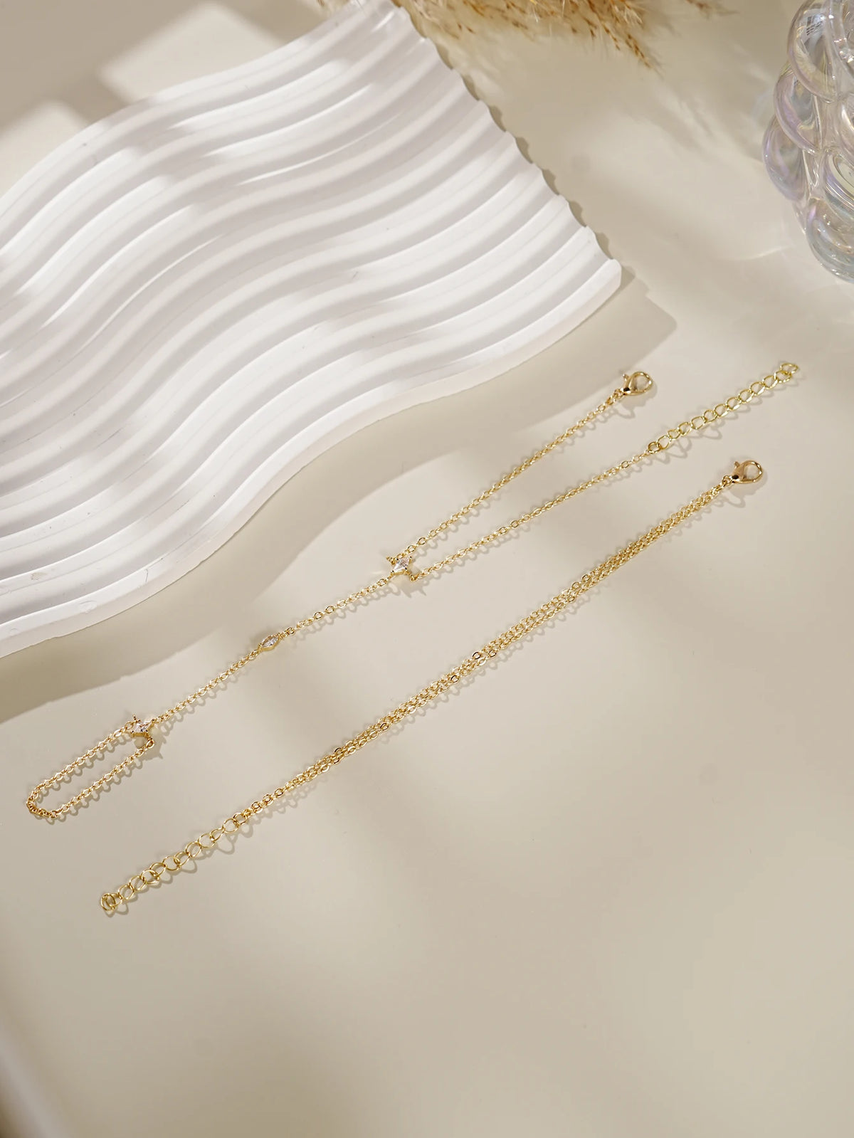 Gold ultra-thin layering chain bracelets displayed on a light background next to a white wavy dish.