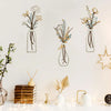 Three glass vases with dried flowers and greenery displayed on a white wall next to stacked wood and small decorative items.