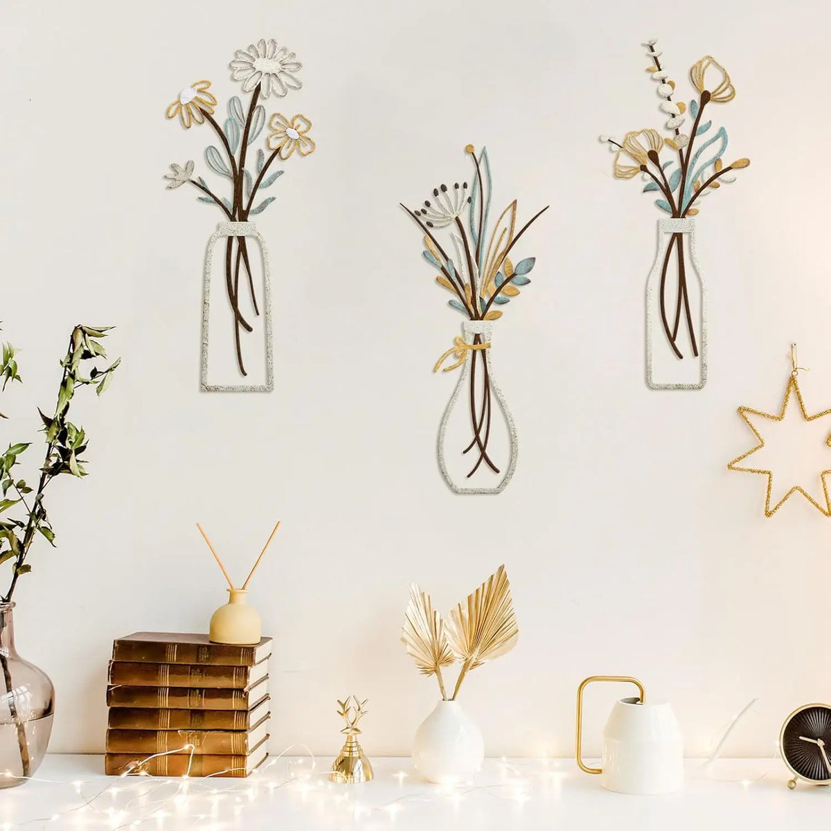 Three glass vases with dried flowers and greenery displayed on a white wall next to stacked wood and small decorative items.