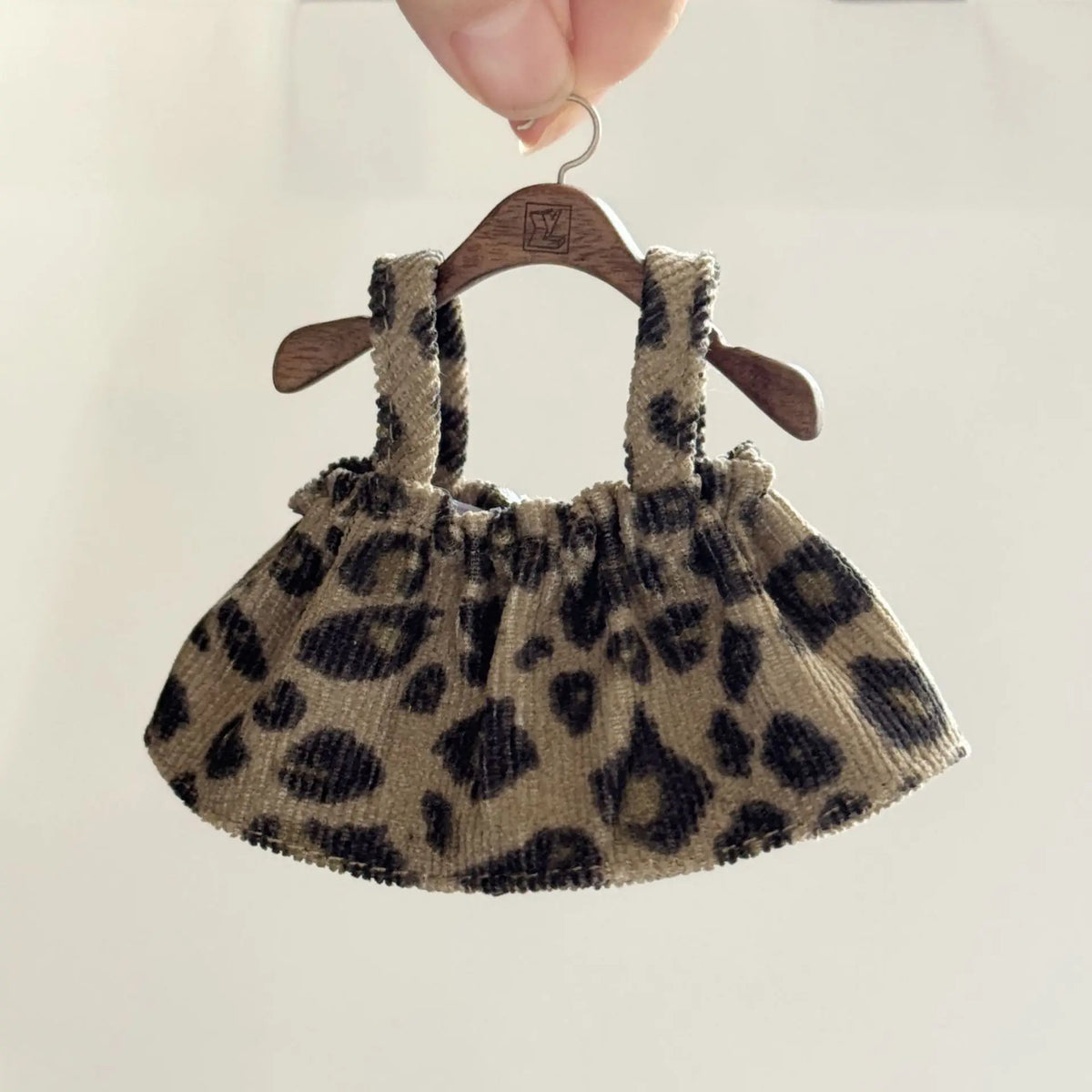 A leopard print shoulder strap skirt for dolls made of brown fabric is displayed on a wooden hanger.