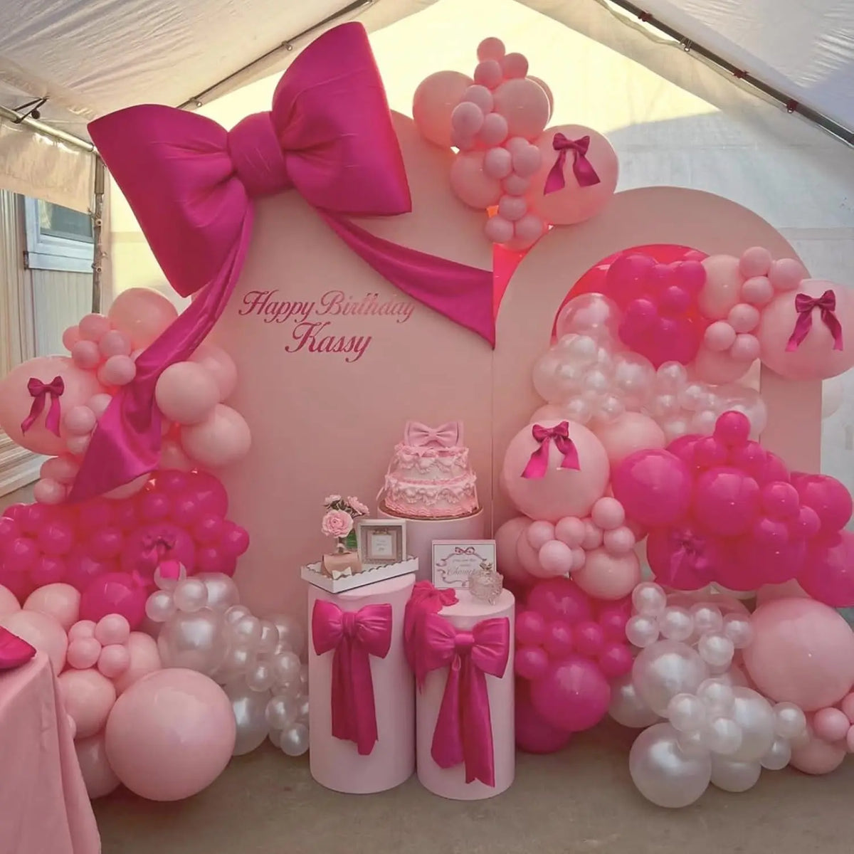 A pink and white balloon party decoration set featuring large balloons and a bow in a tent setting.
