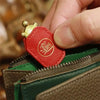 A hand holds a red leather charm shaped like a turtle with a gold head, positioned near a green and brown leather item.