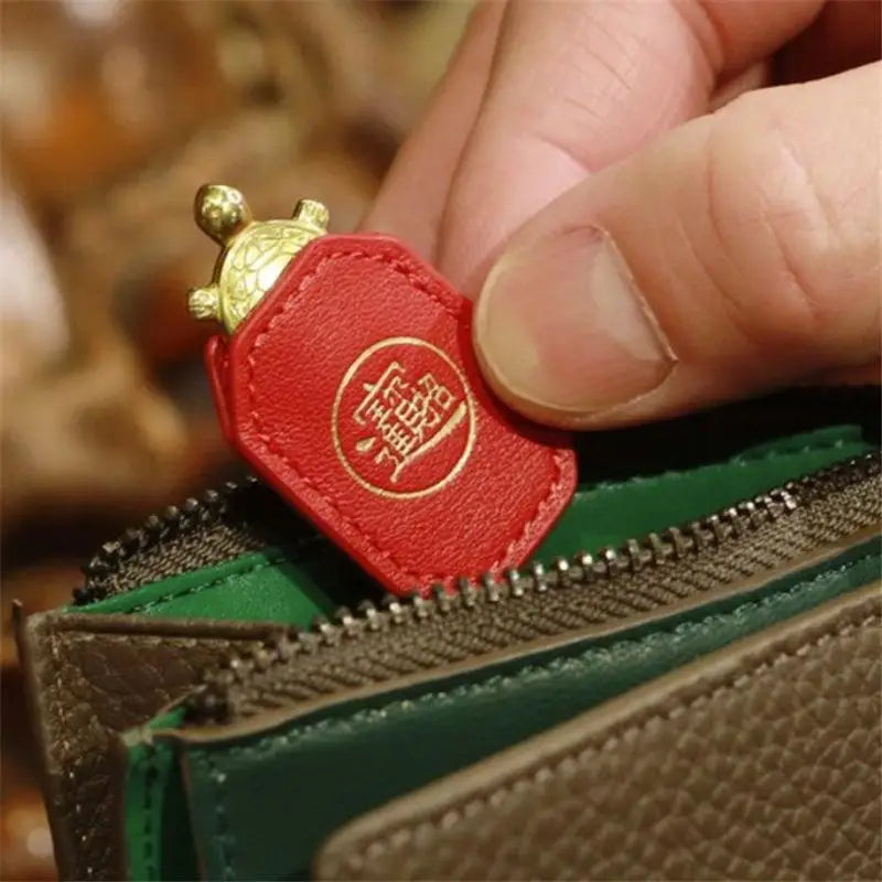 A hand holds a red leather charm shaped like a turtle with a gold head, positioned near a green and brown leather item.