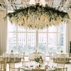 An elegant chandelier made of artificial wisteria flower garlands hangs above a round table in a bright, airy room.