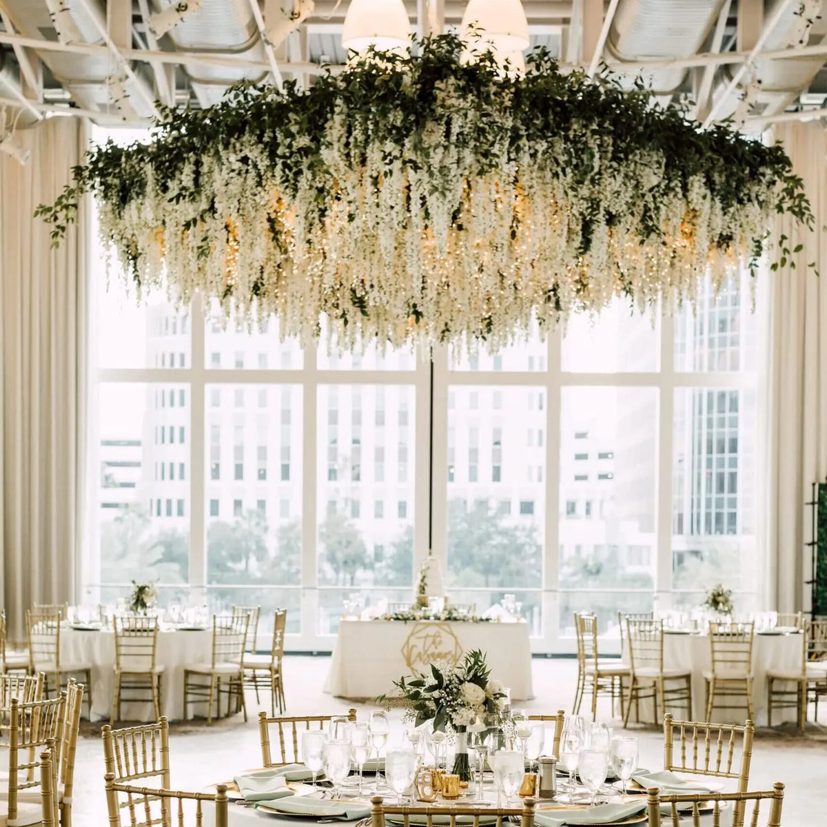 An elegant chandelier made of artificial wisteria flower garlands hangs above a round table in a bright, airy room.