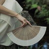 A hand holds a classic bamboo folding fan with decorative pendant in a natural outdoor setting.