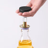 A hand holds a black adjustable bed stabilizer device above a glass bottle containing an amber liquid.