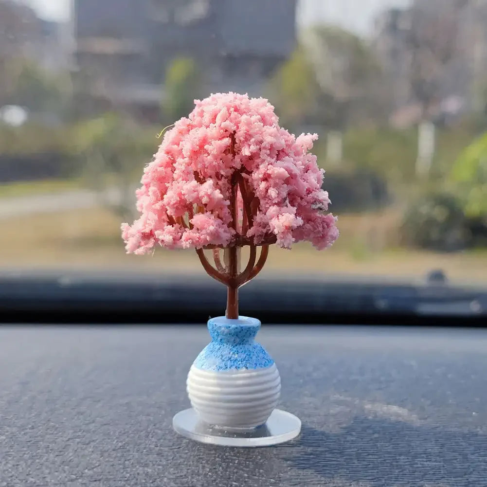 A miniature tree ornament with pink flowers and a blue ceramic base sits on a dark dashboard surface.