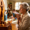 An elderly woman in a beige sweater is praying with folded hands near a wooden statue of the Blessed Mother Mary.