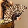 A hand holding a leopard print folding fan with a black frame against a neutral background.