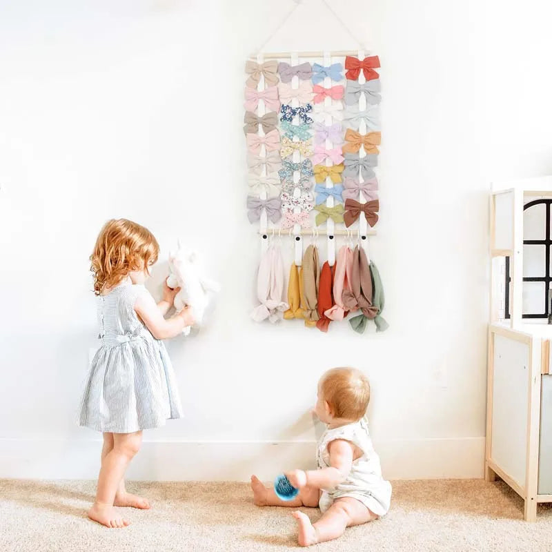 A wall-mounted headband holder for girls with various colored bows displayed on a hanger against a white wall.