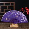 Purple folding fan decorated with white and gold plum blossoms displayed on a wooden stand.
