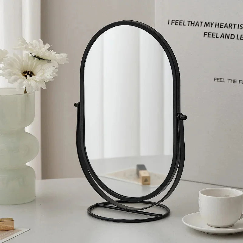 A black oval vanity mirror with a rotating base sits on a white table next to a white cup and a white flower.