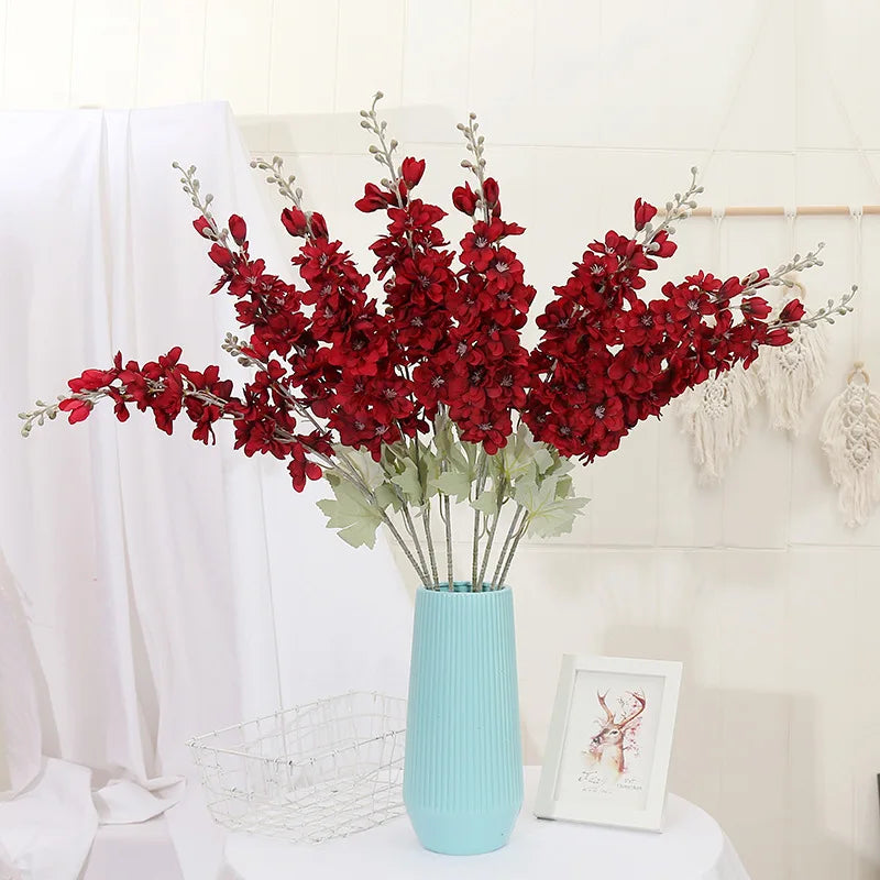 Un bouquet de fleurs artificielles de delphinium rouges dans un vase bleu clair, sur une surface blanche.