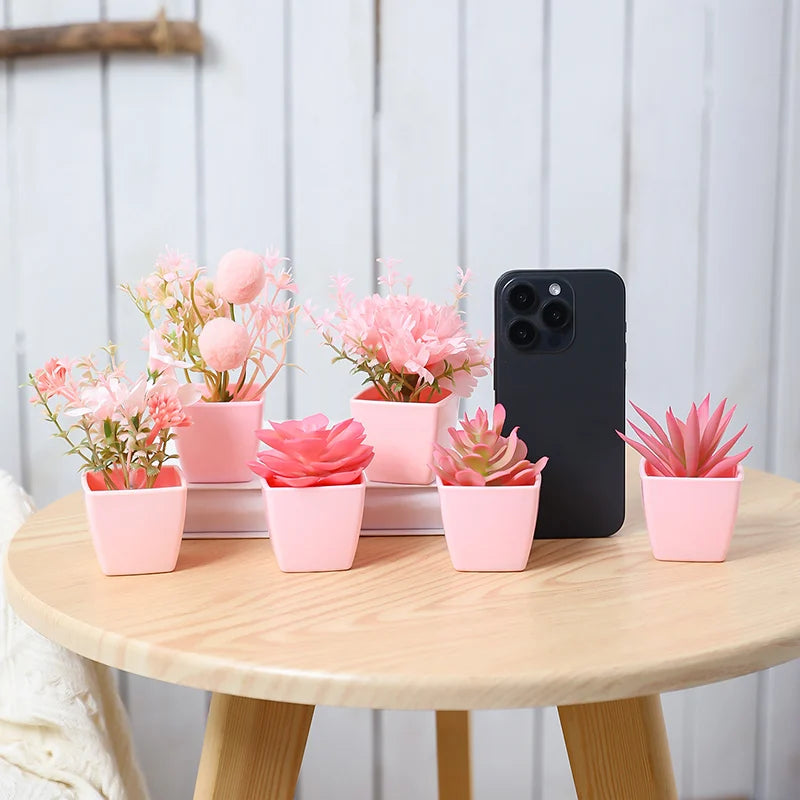 A set of six pink artificial succulent plants in small pots displayed on a wooden table next to a black smartphone.