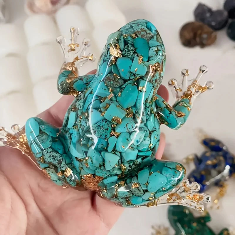 A hand holds a resin craft decoration shaped like a frog with teal and gold crystals.