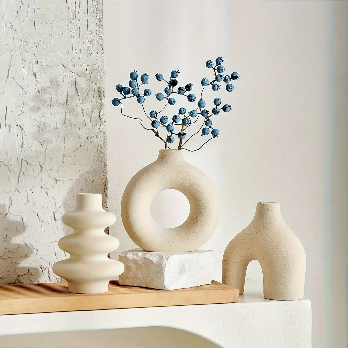 A ceramic vase set featuring a large, smooth, off-white vase with blue berries and a smaller, curved vase on a wooden surface