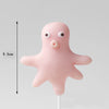 A pink, stylized ocean animal cake topper featuring a cute, round creature with large eyes and a nose, mounted on a white sti