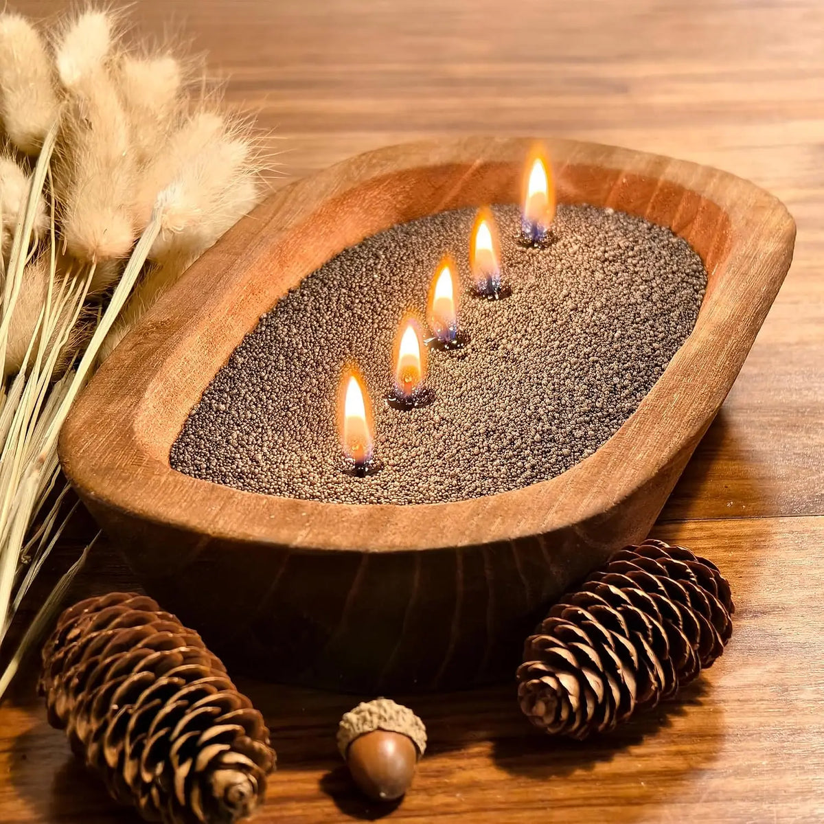 A wooden bowl holds a black pearled candle with wicks, surrounded by pine cones and dried grasses.