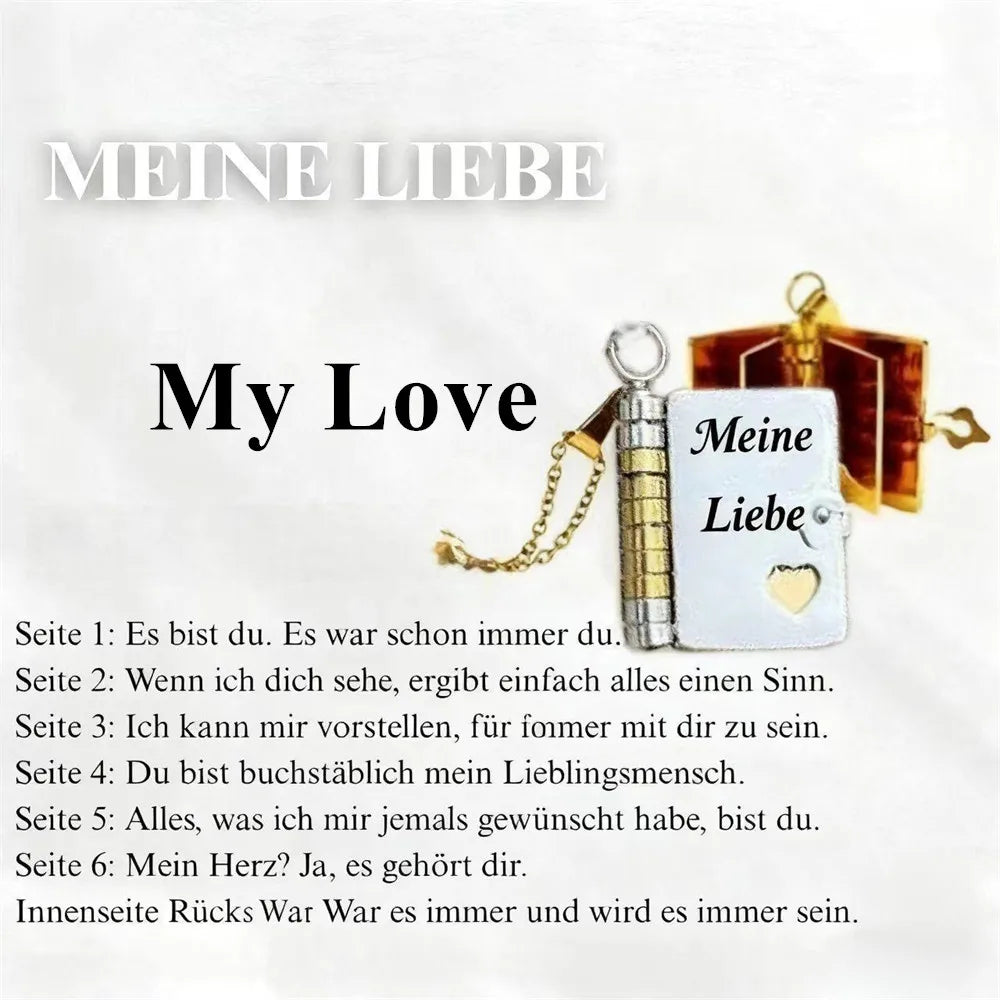 A silver pendant necklace shaped like an open book with a gold clasp and a heart charm, featuring German text.