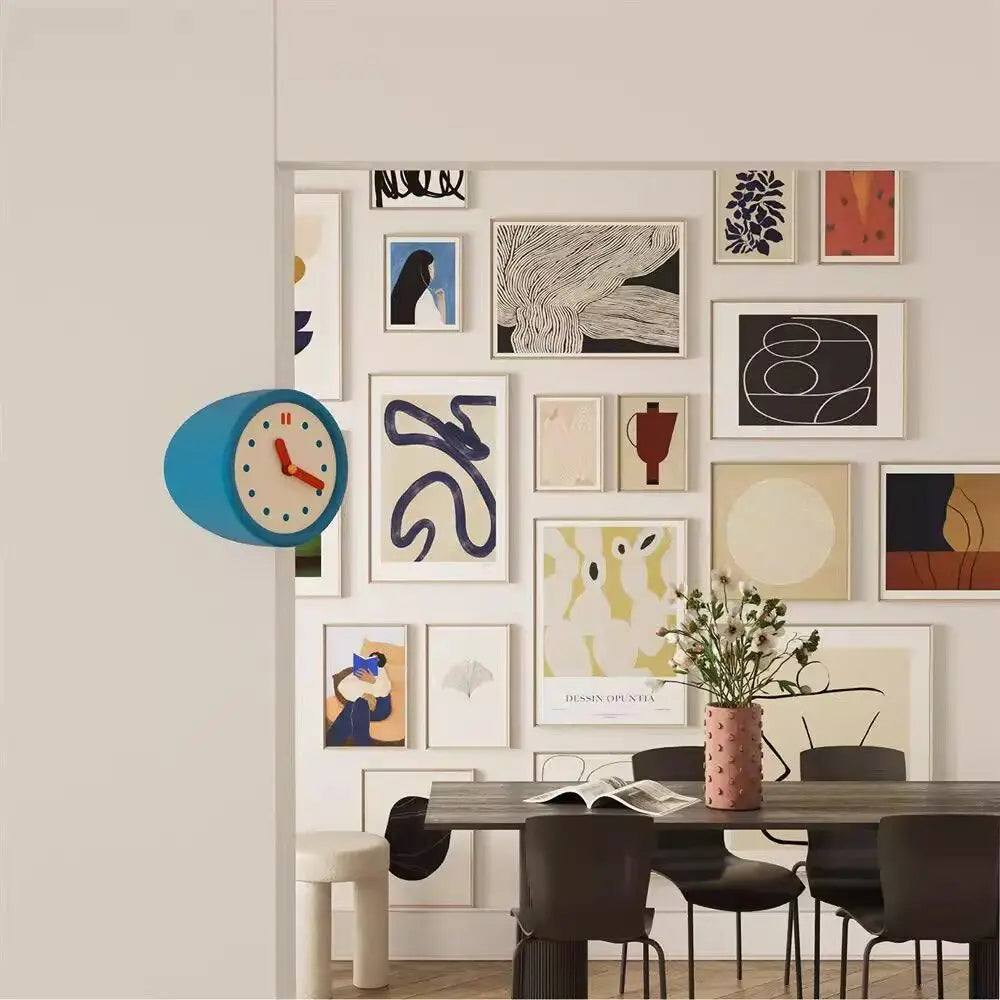 A modern resin wall clock with a blue face and black numbers is displayed on a light wall above various framed artwork.