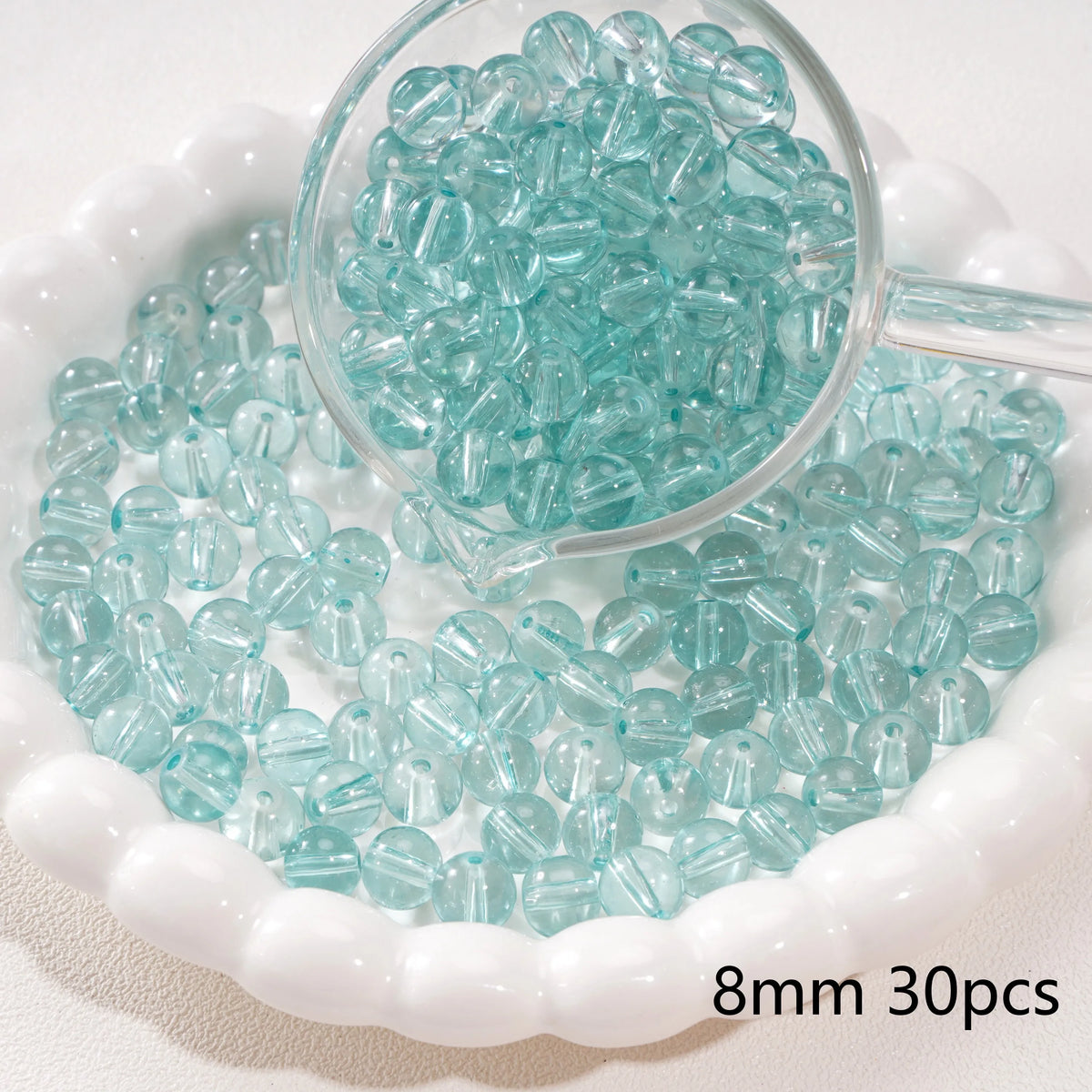 A clear glass measuring spoon filled with small, round, light blue spacer beads resting in a white bowl.