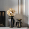 A black ceramic flower vase with gold accents sits on a shelf next to a vertical decorative panel.