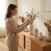 A woman is arranging dried floral branches in a set of modern minimalist matte ceramic vases on a wooden cabinet.