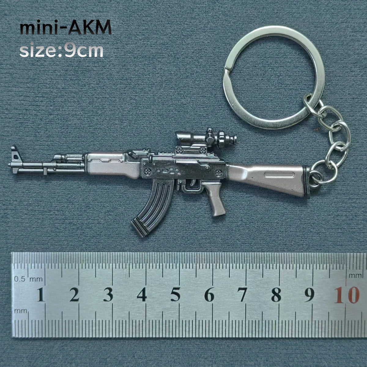 A silver miniature AKM assault rifle model attached to a metal keyring, shown next to a ruler.