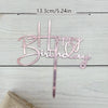 A pink acrylic birthday cake topper with gold lettering spelling out Happy Birthday