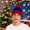 A young man wearing a red knitted beanie with a fluffy pompom and a white shirt, standing in front of a decorated Christmas t