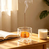 A glass incense burner with a gourd-shaped bowl and a thin stick emitting smoke on a wooden table.