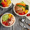 A spread of healthy food including grilled meat, vegetables, and fruit served in disposable paper bowls.