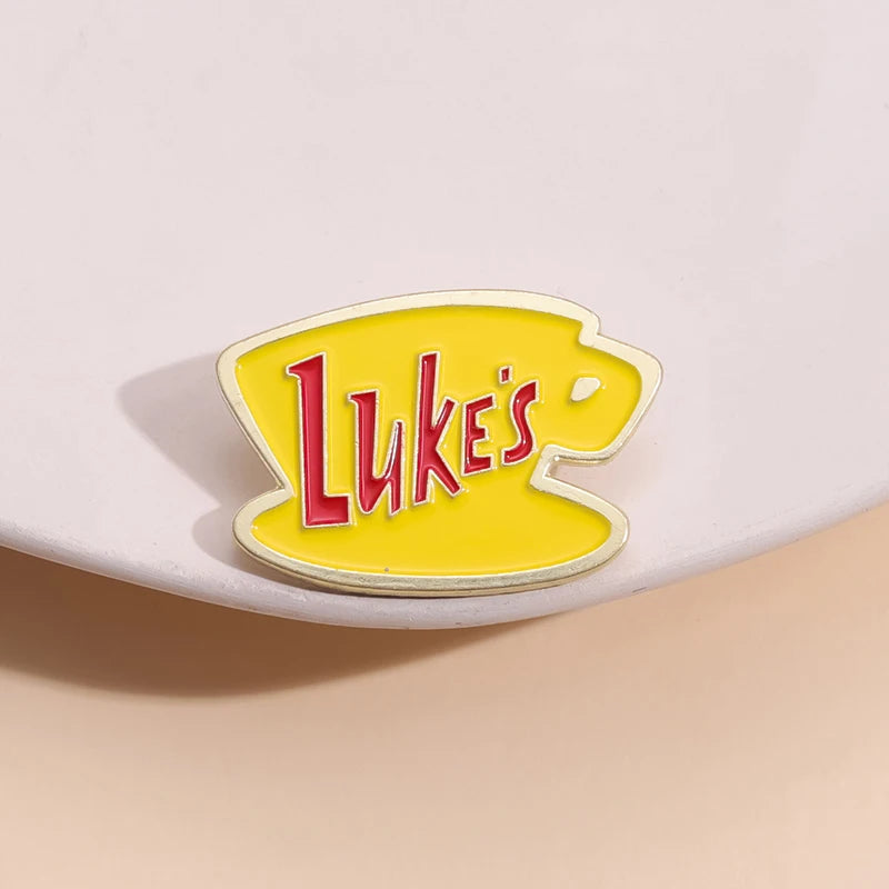 A yellow enamel pin featuring the word Luke's in red text on a white background.