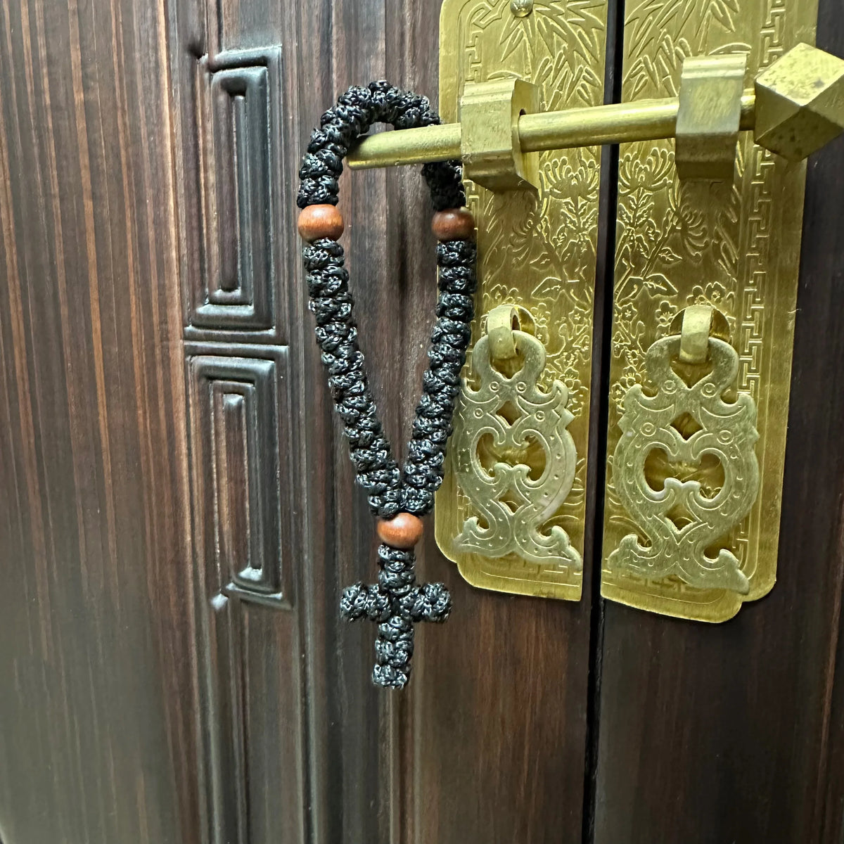 A handmade black prayer rope with a cross charm is displayed on a brass door hanger.