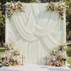 A white satin backdrop curtain with vertical pleats and draped folds decorated with floral arrangements for photography.