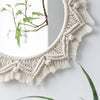 A round macrame wall mirror with a textured, cream-colored fringe hanging on a white wall.