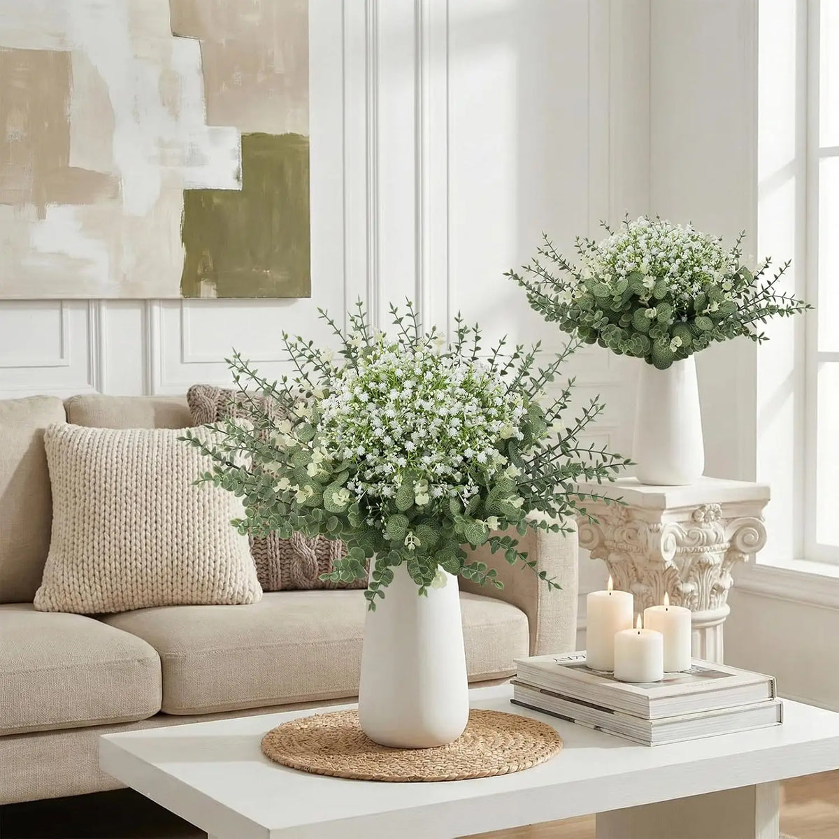 A white ceramic vase filled with baby's breath flowers sits on a white table next to a beige sofa.