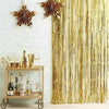 A metallic gold foil tinsel fringe backdrop set hanging on a white brick wall next to a wooden bar shelf.