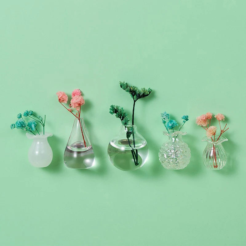 A collection of decorative glass vials and vessels holding dried pink and blue floral sprigs on a light green background.