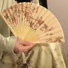 A hand holds an elegant floral bird print bamboo folding fan with pale yellow and brown tones.