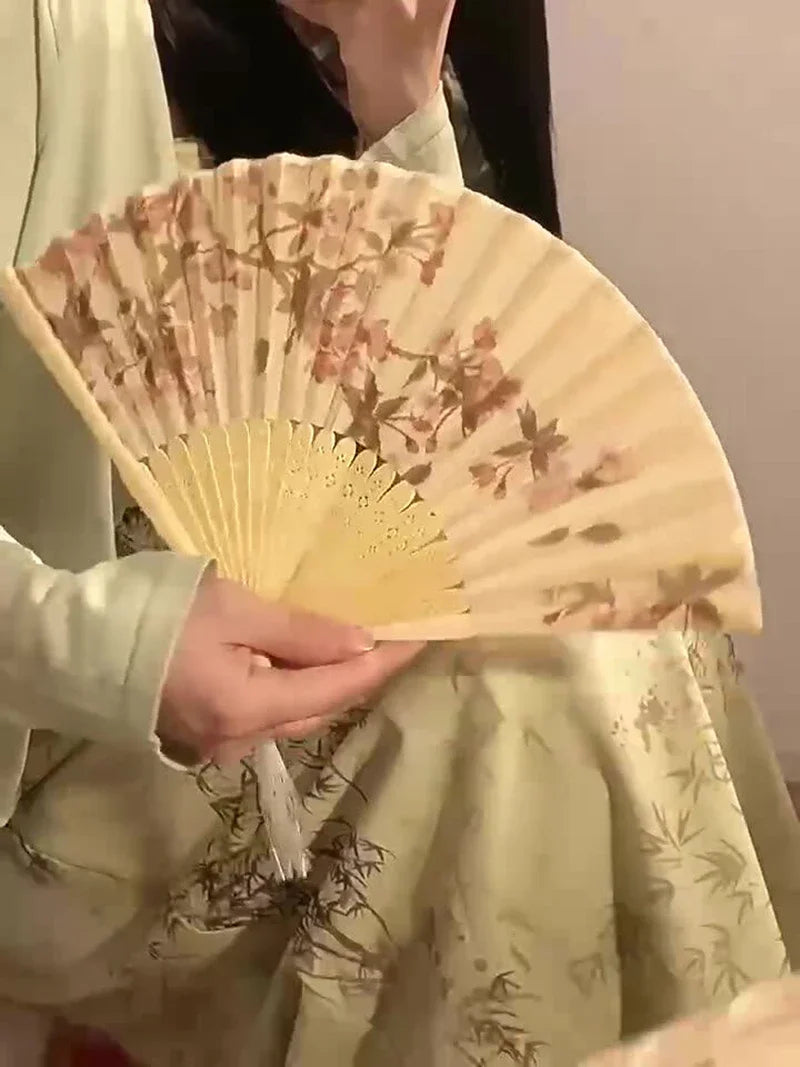 A hand holds an elegant floral bird print bamboo folding fan with pale yellow and brown tones.