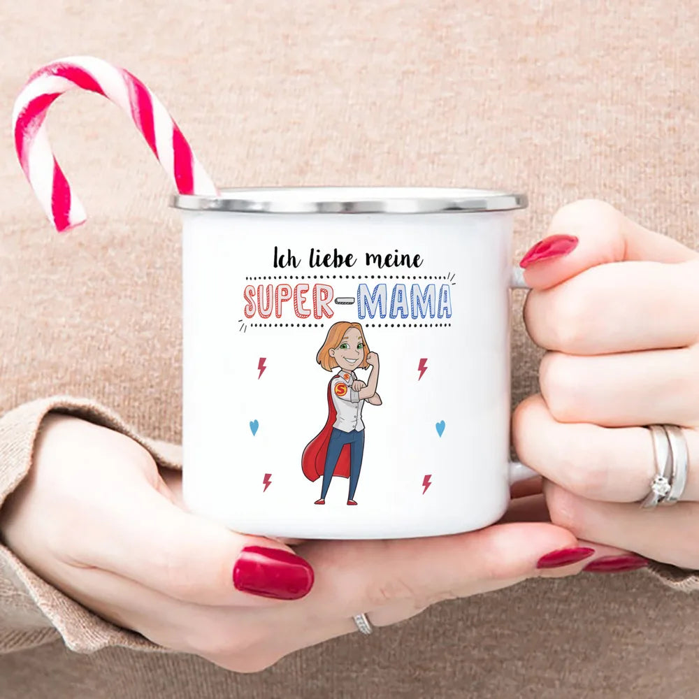 A hand holds a white enamel mug with text reading Ich liebe meine SUPER-MAMA and a red and white striped candy cane.