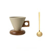 A ceramic coffee mug with a light beige interior and exterior sits on a round wooden base, next to a gold-colored spoon.