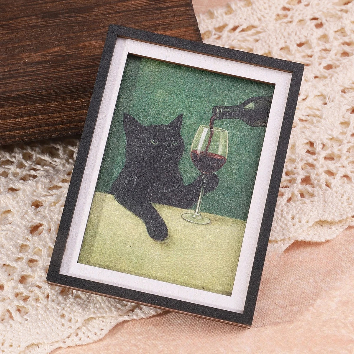 Gerahmtes Foto einer schwarzen Katze, die Rotwein trinkt, vor hellem Hintergrund.
