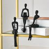 A set of black abstract thinker sculptures displayed on a gold-framed shelf above a stack of books.