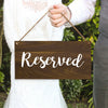 A person holds a rustic wooden sign with the word Reserved written on it, set against a green outdoor background.