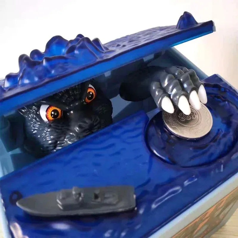 A dark resin Godzilla figure is visible inside a blue piggy bank with an open lid and a coin.