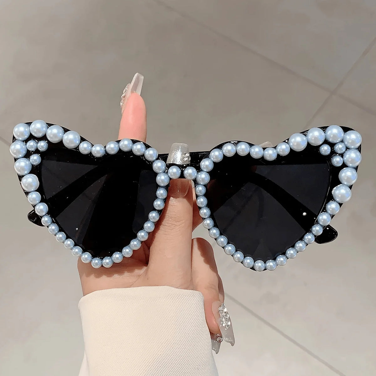 A pair of black heart-shaped sunglasses decorated with white pearls held by a hand.