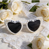 A pair of white pearl-rimmed sunglasses with a heart design and the words bride to be set on a white lace fabric surrounded b
